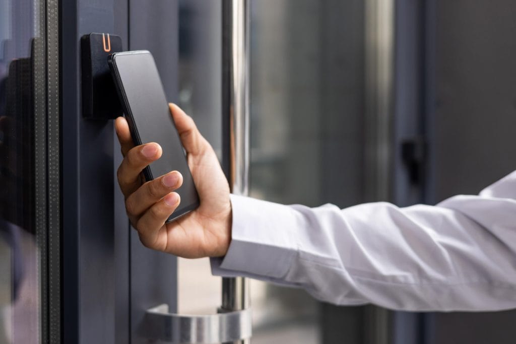 A person in a white shirt holds a smartphone up to an electronic door access panel.