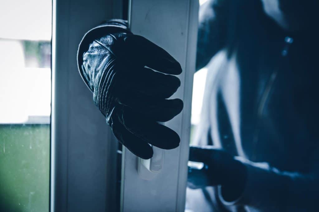 A person in a dark hoodie and gloves is attempting to open a window in a dimly lit setting.