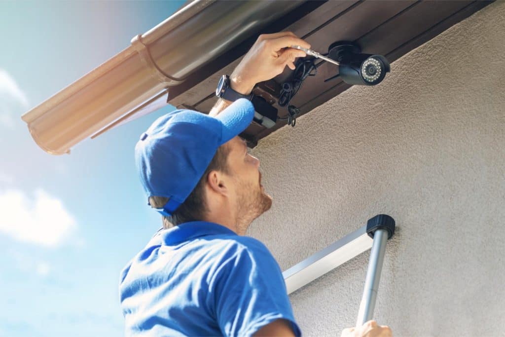 Person in a blue cap and shirt adjusting a security camera under a building's eave, standing on a ladder with a gutter visible.